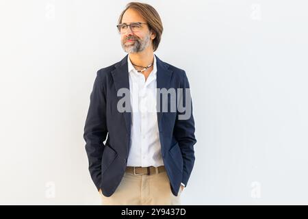 Un uomo maturo con la barba, che indossa occhiali, vestito con un blazer scuro e pantaloni leggeri, si erge con sicurezza su uno sfondo bianco semplice, dall'aspetto Foto Stock
