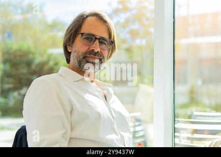 Un uomo d'affari maturo con una camicia bianca e occhiali guarda con sicurezza la fotocamera dal suo ambiente luminoso e moderno, esponendo professionisti Foto Stock