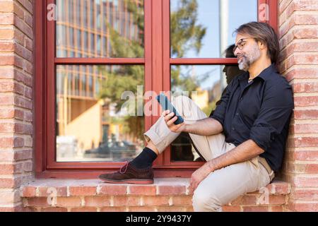 Un uomo d'affari maturo si siede rilassato davanti a una finestra di mattoni, navigando sul suo smartphone in città. Indossa una camicia scura e occhiali casual, godendosi una S. Foto Stock
