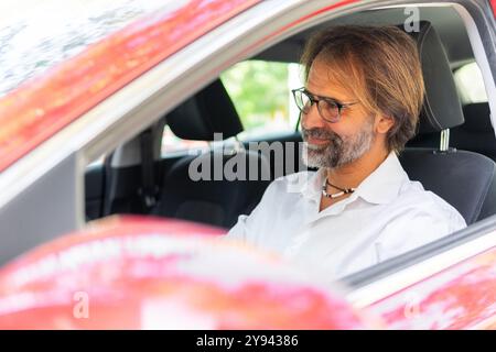 Un uomo di mezza età con barba, occhiali e camicia bianca, guida e guarda lontano dalla telecamera, apparentemente profondamente nel pensiero. Foto Stock