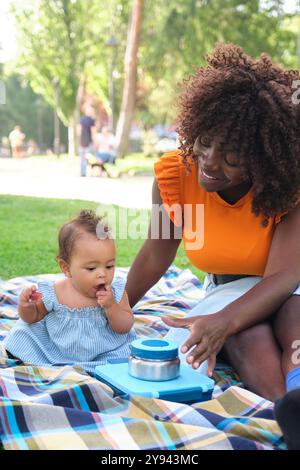 Una allegra madre africana con un top arancione si impegna con la sua bambina su una coperta da picnic in un parco lussureggiante, mettendo in risalto un caldo momento familiare Foto Stock