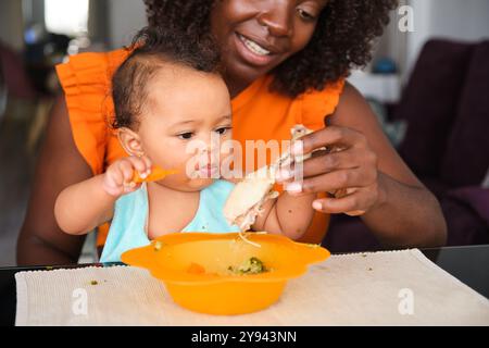 La mamma africana sorridente aiuta la sua bambina a mangiare in un ambiente interno, migliorando i legami di maternità con un momento di nutrizione gioioso catturato dalla fotocamera Foto Stock