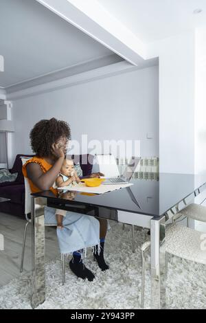 Una mamma africana si impegna a lavorare da casa, seduto su un moderno tavolo di vetro con la sua bambina accanto al suo sorriso riflette una casa calda e appagante Foto Stock