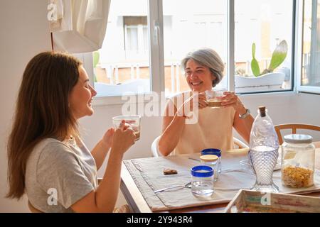 Un momento emozionante immortalato tra madre e figlia, sorseggiando un tè e una conversazione leggera al tavolo della cucina di casa, con illuminazione naturale Foto Stock