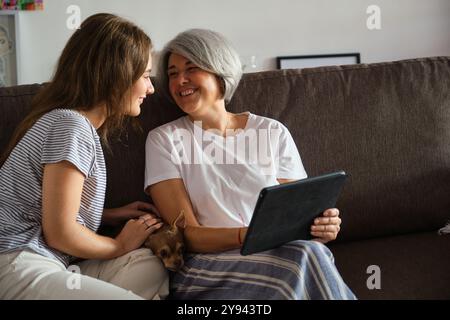 Un momento di grande emozione come madre e figlia adulta condividono una conversazione gioiosa guardando un tablet su un comodo divano, con accanto un piccolo cane Foto Stock