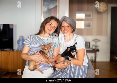 Madre matura felice e figlia adulta, sorridenti e con i loro piccoli cani Chihuahua, comodamente seduti in un ambiente accogliente Foto Stock