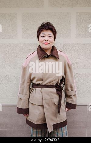 Una donna asiatica di mezza età si erge davanti a una parete beige, vestita con un elegante trench con dettagli marroni, trasudando fiducia e grazia Foto Stock