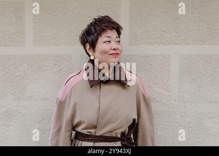 Un'elegante donna di mezza età con capelli corti e rossetto rosso sorride dolcemente, indossando un elegante cappotto rosa e beige su una parete grigia testurizzata Foto Stock