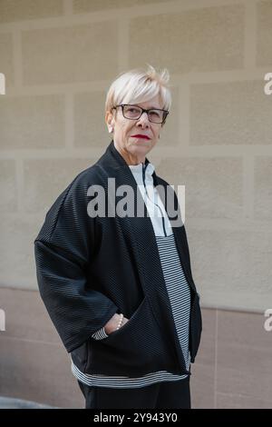 Un'anziana donna in bilico con capelli biondi corti e occhiali, vestita con un'elegante giacca nera testurizzata e camicia a righe, si erge con sicurezza contro un natu Foto Stock