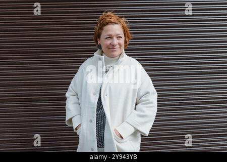 Una donna allegra di mezza età con i capelli rossi corti, che indossa un cappotto bianco e una camicetta a motivi geometrici, posa su uno sfondo a righe scure il suo caldo sorriso Foto Stock