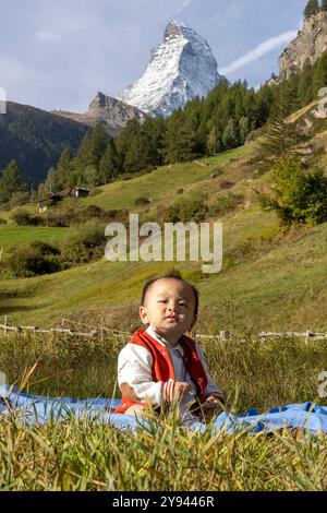 Il bambino coreano vestito con abiti casual siede su una coperta tra le viste panoramiche di Zermatt con l'iconica montagna del Cervino che torreggia nel bac Foto Stock