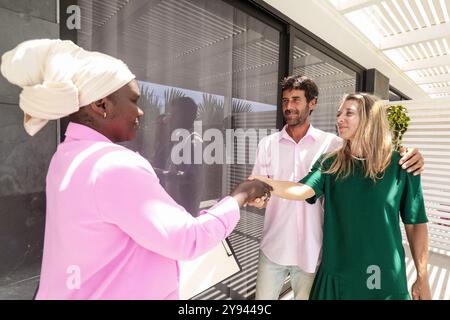 Una donna nera agente immobiliare stringe la mano a una coppia sorridente fuori da una nuova casa, in un patio illuminato dal sole. I clienti, un uomo e una donna, espr Foto Stock