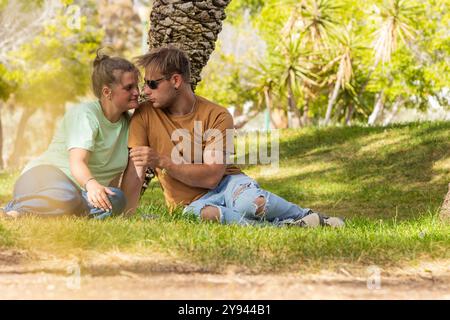 Una scena toccante in un parco illuminato dal sole con una giovane coppia seduta da un albero la donna, sorridente calorosamente, si appoggia verso l'uomo, che ricambia dolcemente la sua af Foto Stock