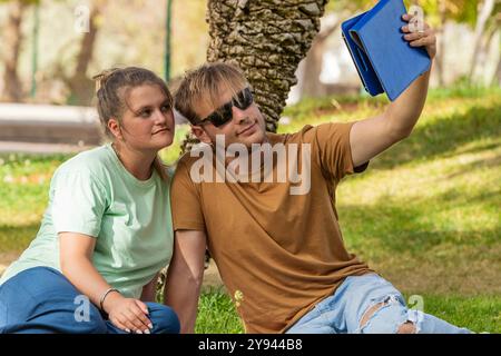 Una giovane coppia si siede sotto un albero ombreggiato in un parco lussureggiante, mentre l'uomo, con gli occhiali da sole, regge un tablet per catturare un momento o mostrare contenuti al suo AT Foto Stock