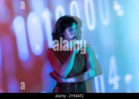 Una giovane donna elegante si staglia con le braccia incrociate sotto luci multicolore, con grandi numeri digitali proiettati sullo sfondo. Foto Stock