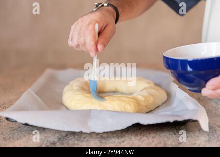 Un primo piano di una mano che applica il lavaggio delle uova all'impasto crudo a forma di anello, utilizzando un pennello blu, per preparare una tradizionale torta dei tre Re Foto Stock