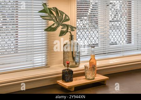 Un elegante ambiente domestico caratterizzato da un elegante vaso di vetro con una singola foglia di monstera, bottiglie decorative e un vetro di cristallo su un vassoio di legno a causa del vento Foto Stock