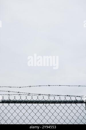 Vista ravvicinata di una recinzione a catena sormontata da filo spinato contro un cielo grigio e nuvoloso. Questa scena urbana cattura il senso di isolamento e sicurezza Foto Stock