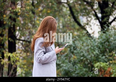 Donna con lunghi capelli di zenzero che indossa un cappotto grigio autunnale in piedi nel parco cittadino con lo smartphone in mano Foto Stock