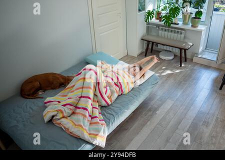 Donna felice che sdraia a letto con il cane risvegliato dal sonno sano lentamente svegliandosi allungando le braccia. Foto Stock