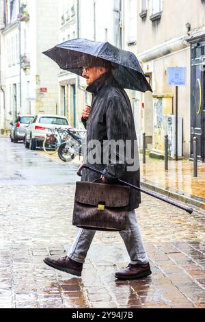 Uomo d'affari anziano con valigetta, bastone da passeggio e ombrello che cammina lungo la strada della città sotto la pioggia battente - Tours, Indre-et-Loire (37), Francia. Foto Stock