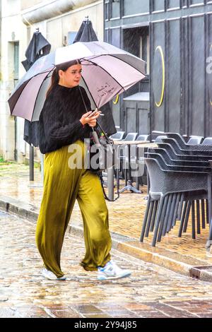 Donna in pantaloni larghi con ombrello che cammina lungo la strada del centro città sotto la pioggia battente - Tours, Indre-et-Loire (37), Francia. Foto Stock