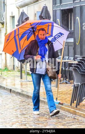 Donna con ombrello pubblicitario che cammina lungo la strada del centro città sotto la pioggia battente - Tours, Indre-et-Loire (37), Francia. Foto Stock