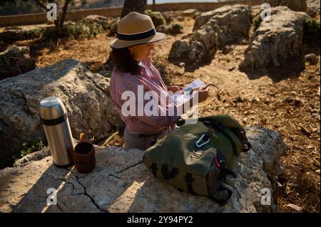 Una donna che indossa un cappello legge una mappa mentre siede sulle rocce in un ambiente naturale e soleggiato. Uno zaino e thermos si siedono accanto a lei, suggerendo una giornata di adven Foto Stock