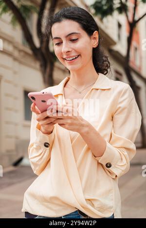 Ritratto verticale di una giovane donna adulta sorridente che usa un cellulare all'aperto, in piedi in un'area urbana mentre interagisce con il suo smartphone, concentrata e impegnata, trascorrendo una giornata all'aperto. Foto di alta qualità Foto Stock