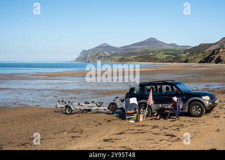 Una donna si rilassa su una sdraio accanto a un SUV sulla spiaggia di Nefyn sulla penisola di Llŷn nel Galles del Nord, con la collina di Yr Eifl sullo sfondo. Foto Stock
