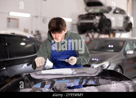 Giovane tecnico della carrozzeria che applica il riempitivo al paraurti dell'auto danneggiato in garage Foto Stock