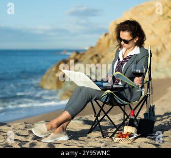 Donna professionista in tuta che lavora a distanza e beve vino in spiaggia Foto Stock