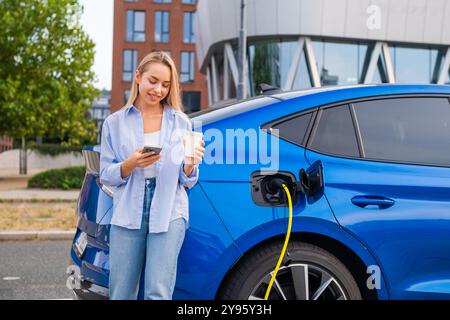 Una giovane donna che ricarica la sua auto elettrica mentre si gusta un caffè e usa il suo smartphone per pagare in strada Foto Stock