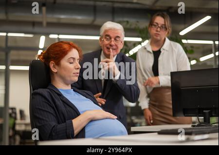 Anziano uomo e donna caucasica che riscuotono dietro il loro collega incinta addormentato in ufficio. Foto Stock