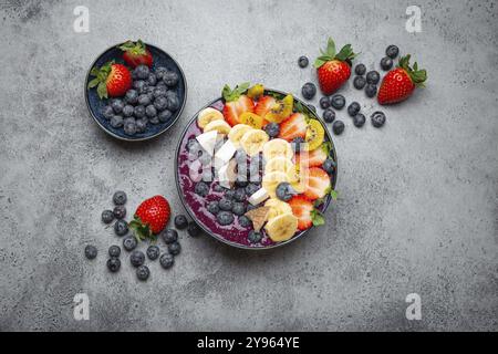 Frullato estivo di acai salutare con semi di chia, banana fresca, fragole, mirtilli, cocco, vista dall'alto kiwi su sfondo rustico in cemento con cucchiaio Foto Stock