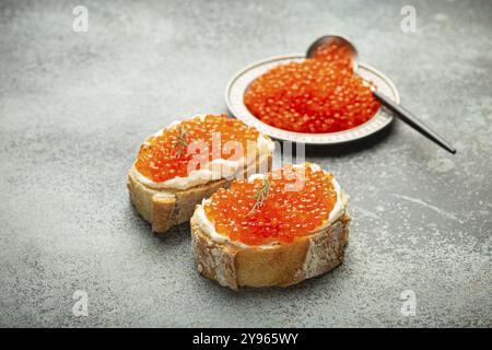 Piccolo piatto di metallo con caviale di salmone rosso e due crostini di caviale su sfondo grigio di cemento, prelibatezze e antipasti di lusso, fotogr Foto Stock