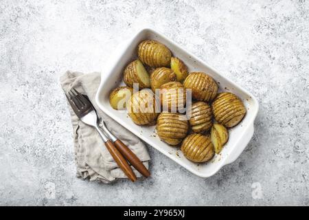 Patate Hasselback alle erbe in casseruola bianca in ceramica su fondo bianco tavolo di cemento dall'alto. Deliziosa potata fatta in casa a base di fisarmonica Foto Stock