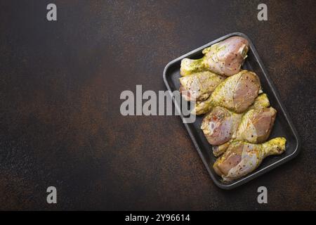 Cosce di pollo crude non cotte in marinata verde con condimenti in contenitore di plastica nera vista dall'alto su sfondo rustico scuro. Preparazione di un pasto sano con Foto Stock