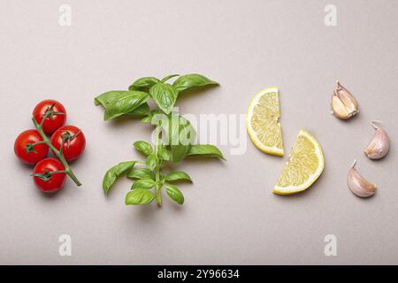 Composizione con ingredienti alimentari sani: Ramo di pomodori ciliegini freschi, ramo di basilico, spicchi d'aglio, spicchi di limone su dorso pulito grigio minimalista Foto Stock