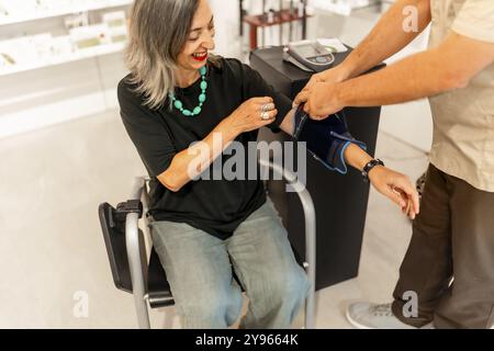 Donna matura e felice seduta su una sedia in farmacia per controllare la pressione sanguigna Foto Stock