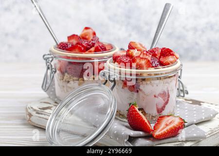 Parfait con fragole fresche, yogurt e granola croccante in vasetti di vetro trasparente su sfondo bianco rustico in legno da angolo, salute Foto Stock