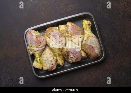 Cosce di pollo crude non cotte in marinata verde con condimenti in contenitore di plastica nera vista dall'alto su sfondo rustico scuro. Preparazione di un pasto sano con Foto Stock