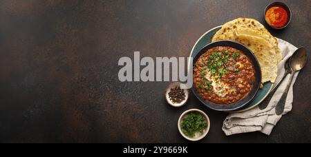 Tradizionale piatto punjabi indiano dal makhani con lenticchie e fagioli in ciotola nera servito con pane piatto naan, coriandolo fresco e due cucchiai di pane marrone Foto Stock