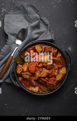 Stufato di carne di manzo con patate, carota e delizioso sugo in casseruola nera con foglie di alloro con cucchiaio su fondo nero scuro rustico in cemento fr Foto Stock