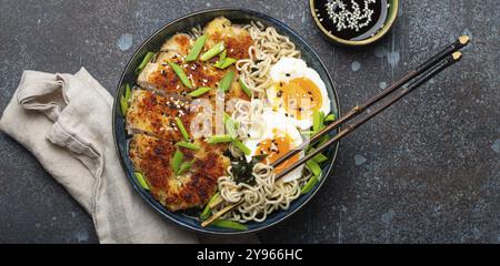 Zuppa di ramen di pasta asiatica con filetto di pollo fritto al panko e uova sode in un recipiente di ceramica con bastoncini di tritare e salsa di soia su backgroun rustico di pietra Foto Stock