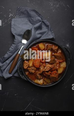 Stufato di carne di manzo con patate, carota e delizioso sugo in casseruola nera con foglie di alloro con cucchiaio su fondo nero scuro rustico in cemento fr Foto Stock