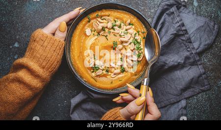 Mani femminili in maglione a maglia giallo che tiene una ciotola con zuppa di crema di zucca su fondo di pietra scura con cucchiaio decorato con arachidi, vista dall'alto. Au Foto Stock