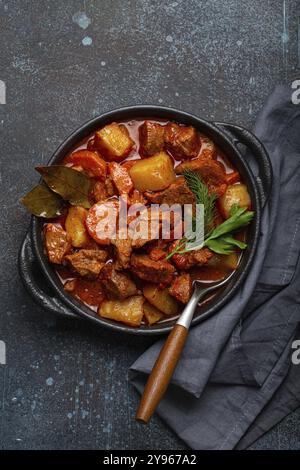 Stufato di carne di manzo con patate, carota e delizioso sugo in casseruola nera con foglie di alloro e erbe fresche verdi con cucchiaio su nero scuro rustico Foto Stock