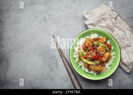 Pollo asiatico agrodolce e appiccicoso con verdure, friggere e riso in una ciotola di ceramica con bacchette, vista dall'alto su fondo in pietra rustica grigia, mestiere Foto Stock