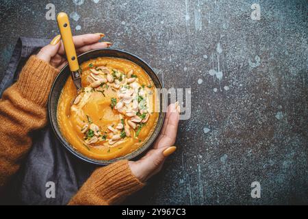 Mani femminili in maglione a maglia giallo che tiene una ciotola con zuppa di crema di zucca su fondo di pietra scura con cucchiaio decorato con arachidi, vista dall'alto. Au Foto Stock